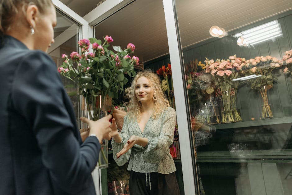 A woman with curly blonde hair, wearing a light-colored floral blouse, is standing inside a flower shop at night, smiling as she receives a bouquet of vibrant pink roses with green foliage from a flow