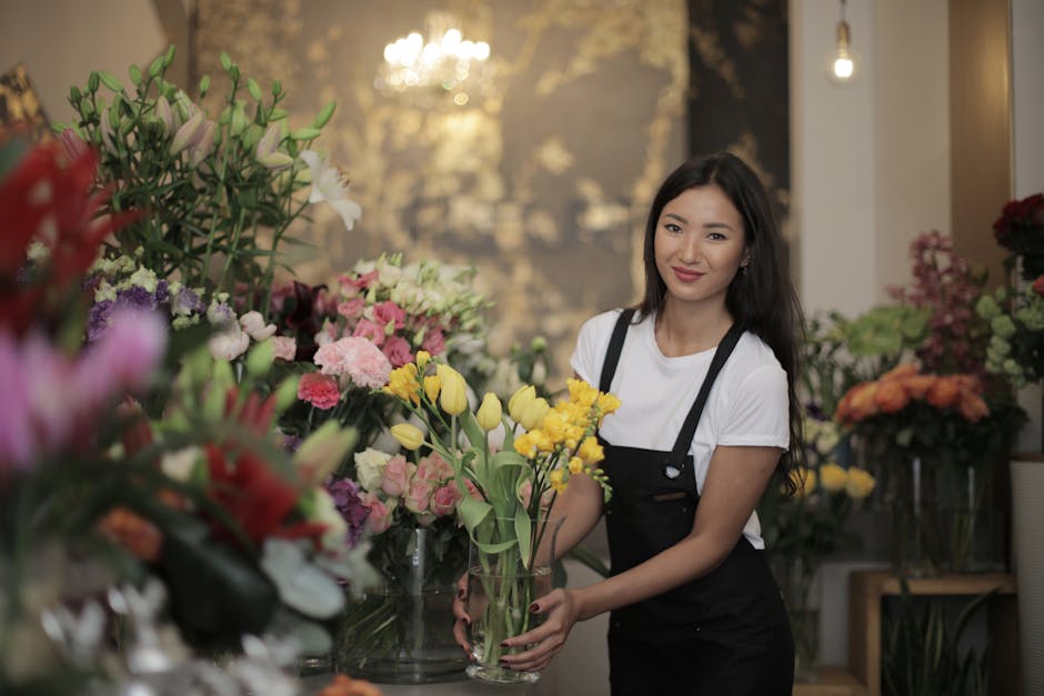 A young woman with long, dark hair, wearing a white t-shirt and a black apron, is holding a clear glass vase filled with yellow tulips and greenery inside a florist shop. The background features a var