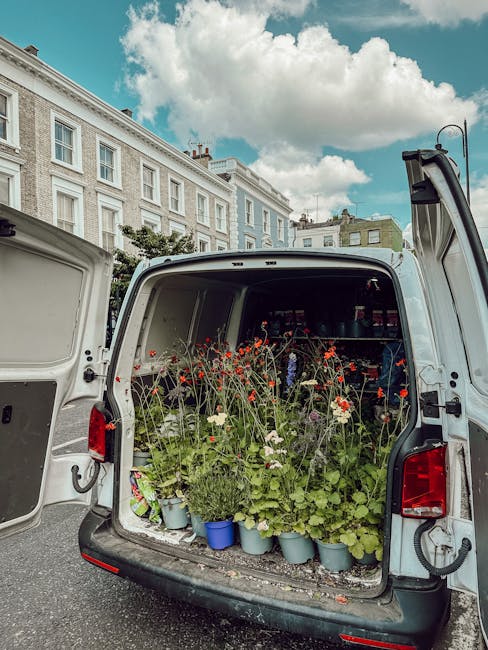 The open back of a white delivery van filled with potted flowers, including red poppies, white daisies, and green foliage, arranged neatly on the vehicle’s floor and shelves. The flowers appear fres