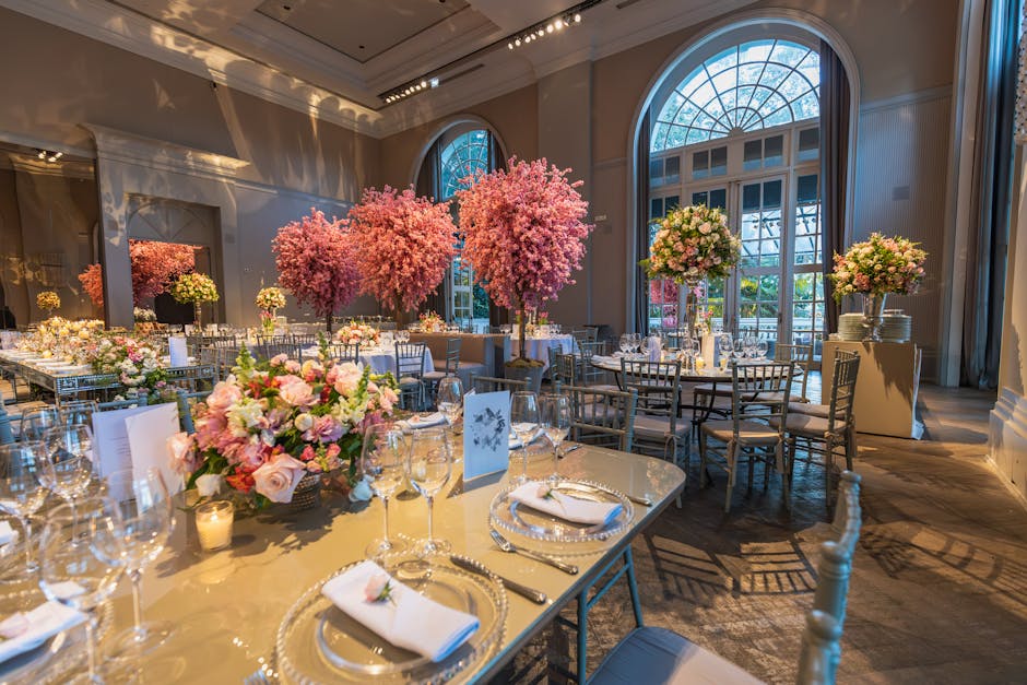 The image shows an elegant indoor event space decorated with floral arrangements and cherry blossom trees. The tables are set with fine glassware, silver cutlery, and neatly folded white napkins, crea