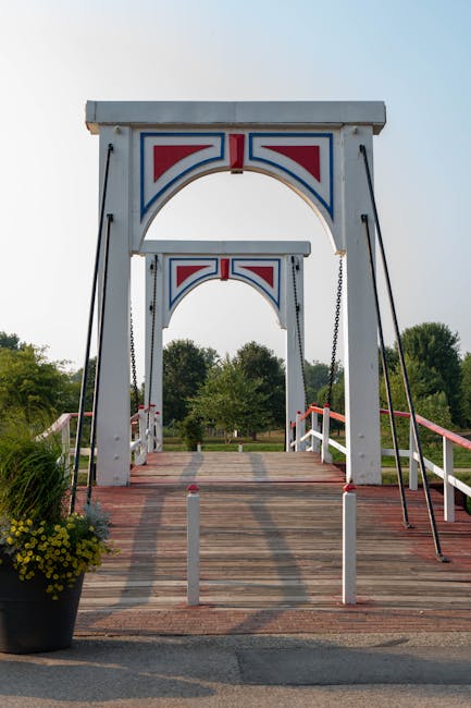 A small decorative bridge with a white wooden structure accented by red, blue, and white Union Jack-style motifs on the arched top sections, supported by black metal chains on either side. The bridge 