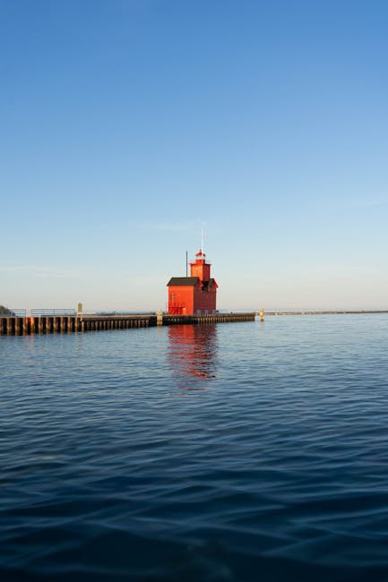 The image features a serene scene of a red lighthouse situated at the end of a wooden pier extending into calm, reflective water. The lighthouse has a rectangular shape with a small turret on top and 