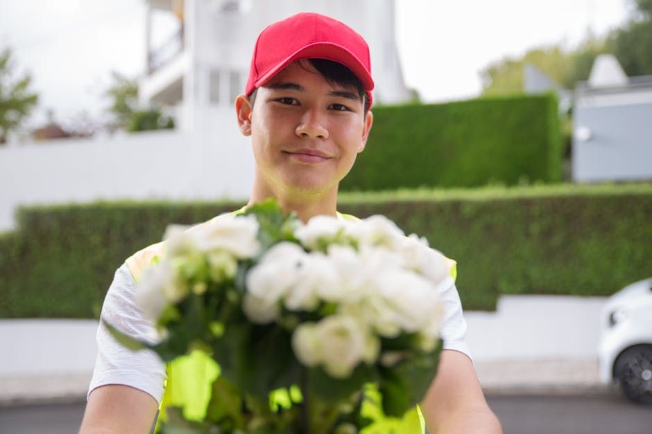 A delivery person wearing a red cap and a reflective yellow vest is holding a bouquet of white flowers with green foliage, extending it towards the camera. The bouquet appears fresh, with delicate pet