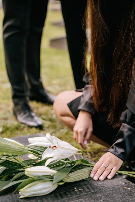 A floral arrangement of white lilies with large, trumpet-shaped petals and prominent stamens lying on a dark stone surface. The lilies are fresh, with glossy green leaves and long, sturdy stems. A wom
