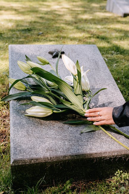 A bouquet of white lilies with large, elegant petals and long green stems, arranged on a flat, polished grey granite grave marker. The flowers are freshly cut with vibrant, healthy leaves, and some ar